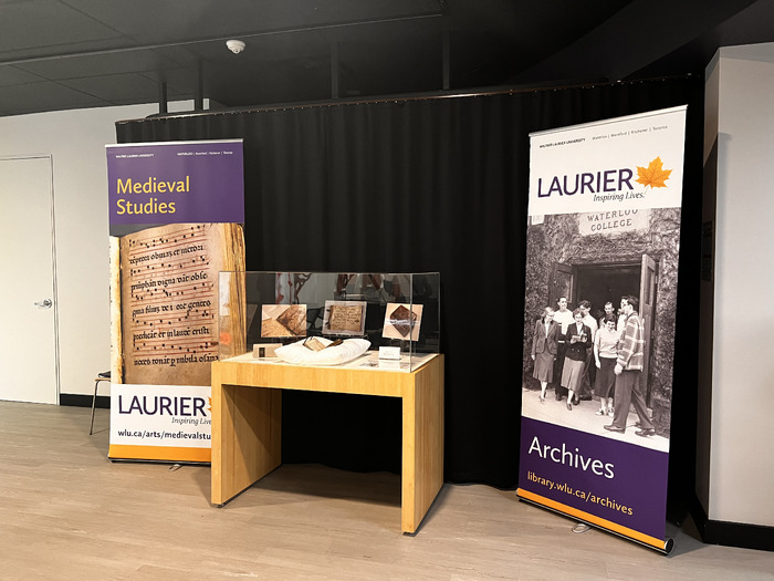 Display of the Noted Hymnal showcased in a glass display case, in between two banners representing the Laurier Archives and the Medieval Studies department.