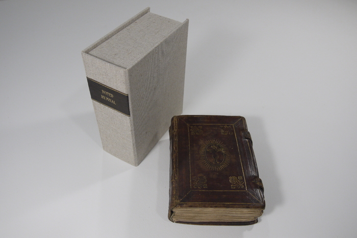 The Noted Hymnal is laid on a surface beside the custom made box which has a label on it that reads "Noted Hymnal".